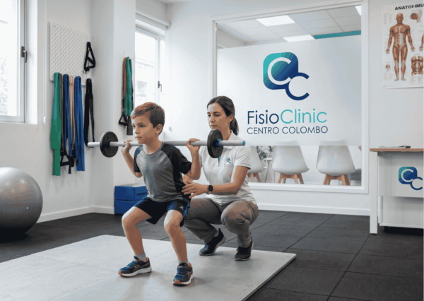 Fisioterapista della FisioClinic Centro Colombo Genova guida un bambino in età evolutiva nell’esecuzione di squat con bilanciere durante una seduta di fisioterapia pediatrica in palestra riabilitativa